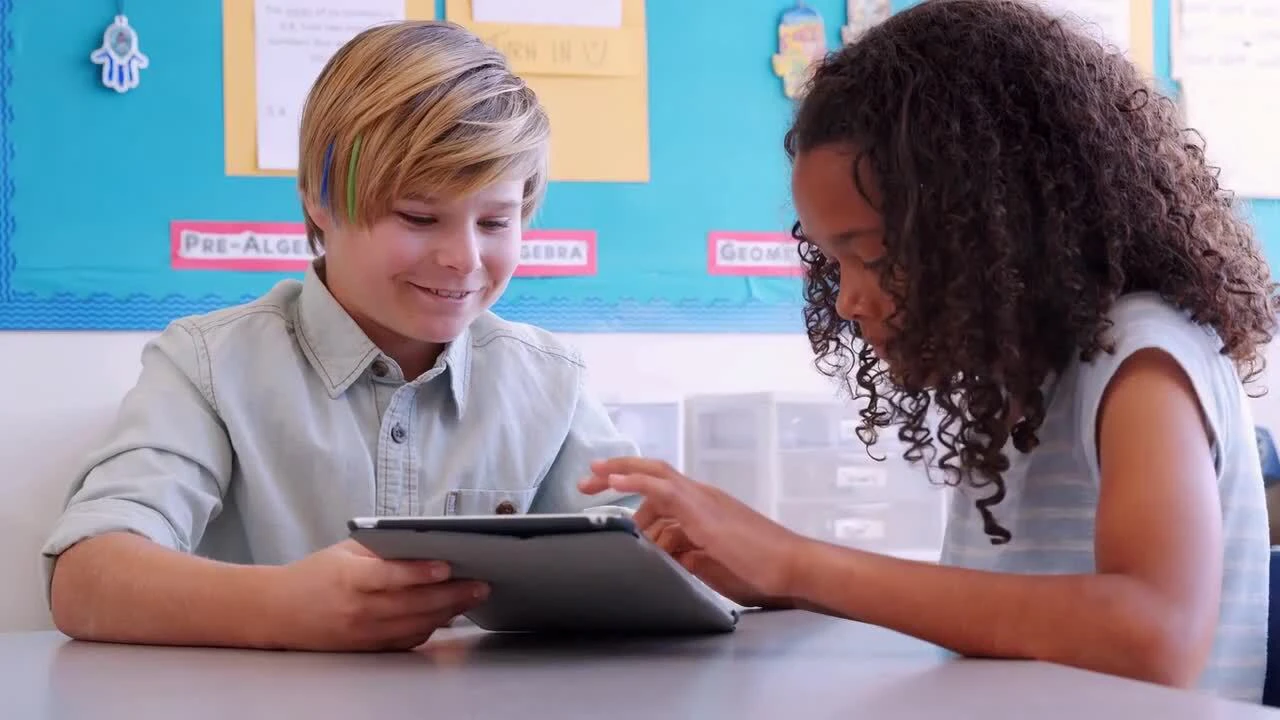 Two children using a tablet together at a desk in a classroom. A bulletin board with educational content is in the background.